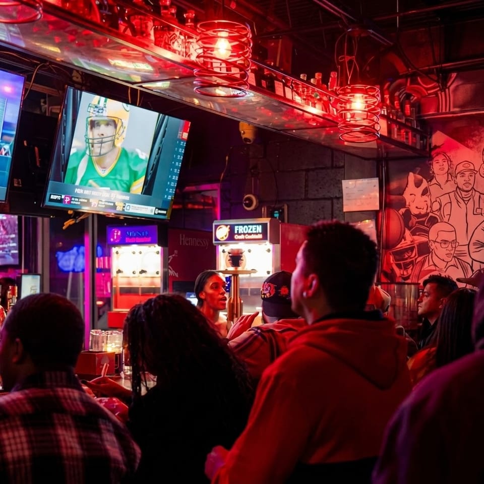 Crowded sports bar glowing with red lighting, showing a football game on television, frozen drink machines, and mural artwork.
