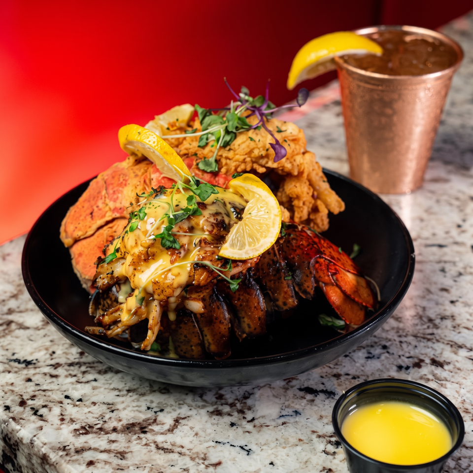 Lavish seafood platter with lobster, shrimp, grilled shellfish, crispy sides, lemon wedges, herbs, and dipping sauces on a black plate.