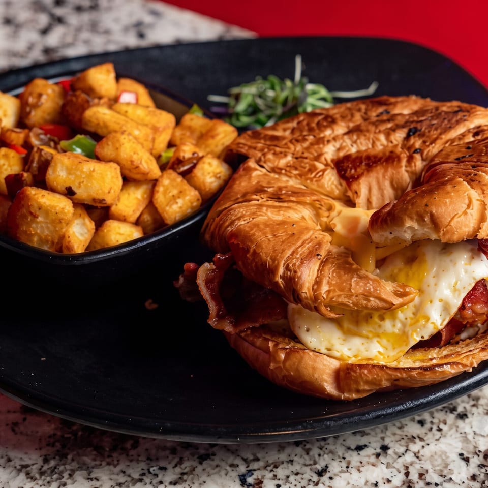 Croissant sandwich with bacon, egg, and melted cheese served alongside seasoned roasted potatoes on a black plate.
