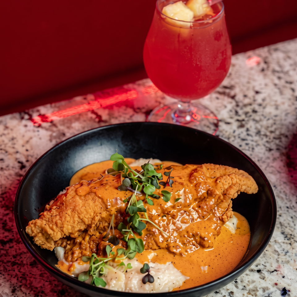 Crispy fried chicken topped with creamy orange sauce and microgreens, served in a black bowl beside a red fruit cocktail.
