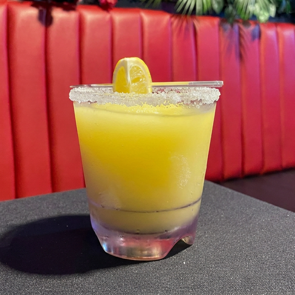 Yellow citrus cocktail in a sugar-rimmed glass with a lemon garnish on a restaurant table, against red upholstered seating.