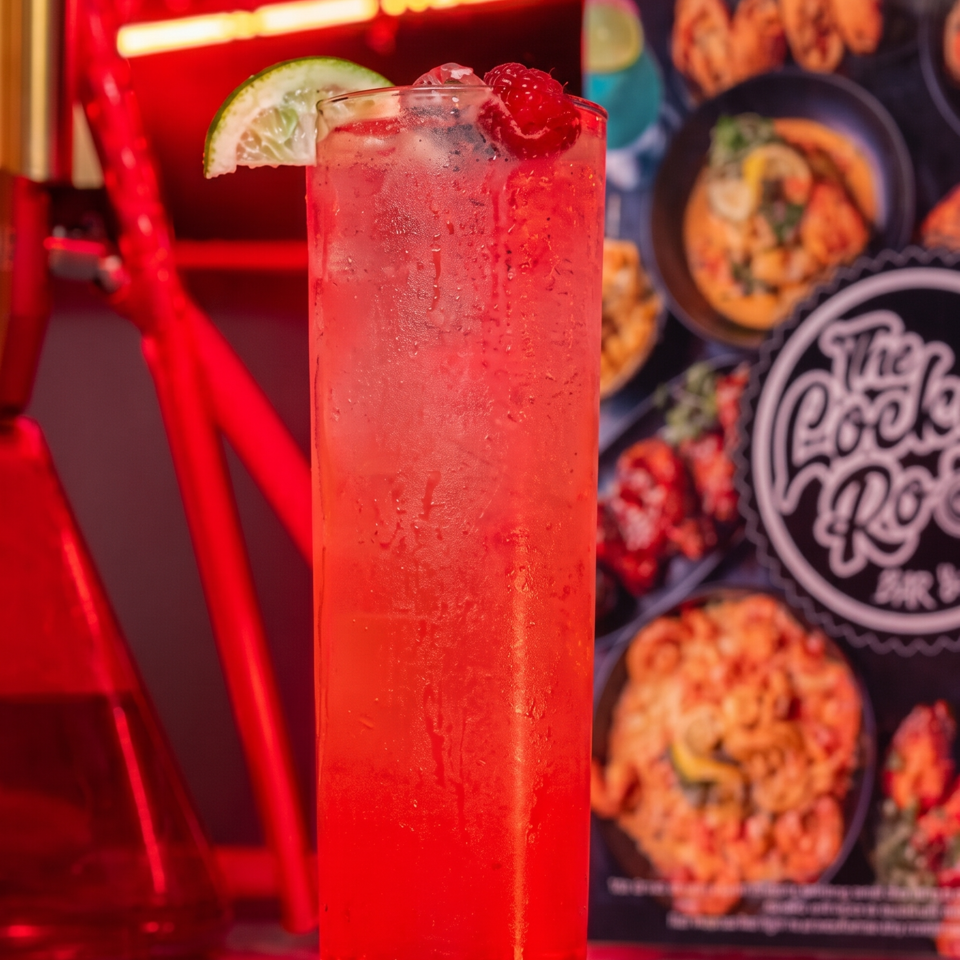 Tall chilled red fruit drink garnished with lime and raspberry, set against a vibrant restaurant backdrop.