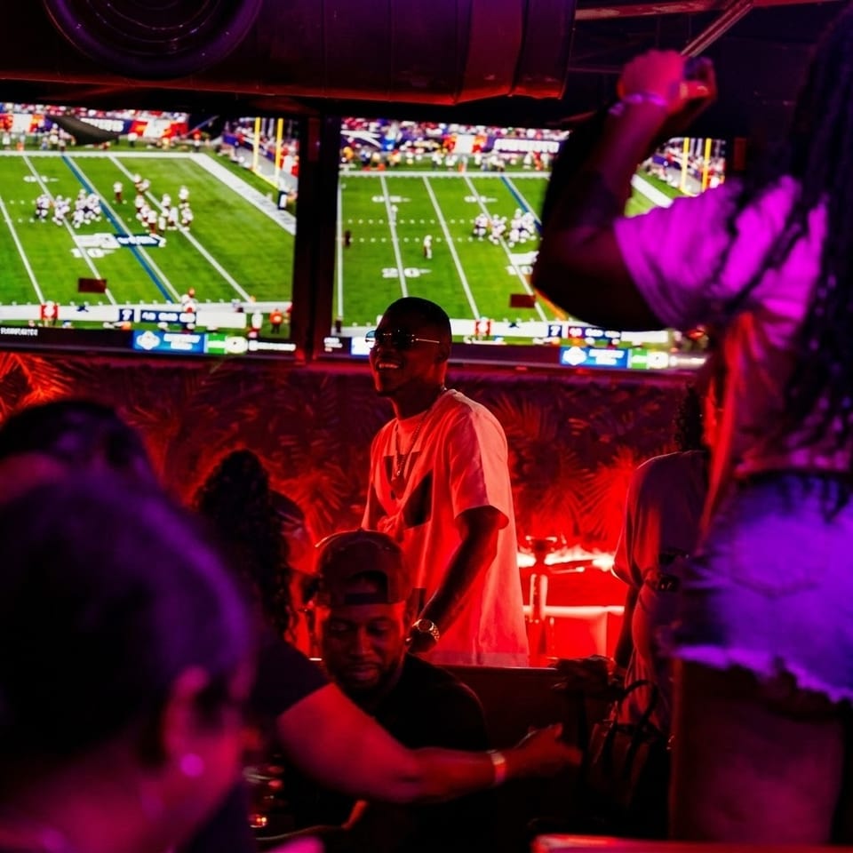 Crowded sports bar bathed in purple and red light, with patrons watching football on multiple large screens.