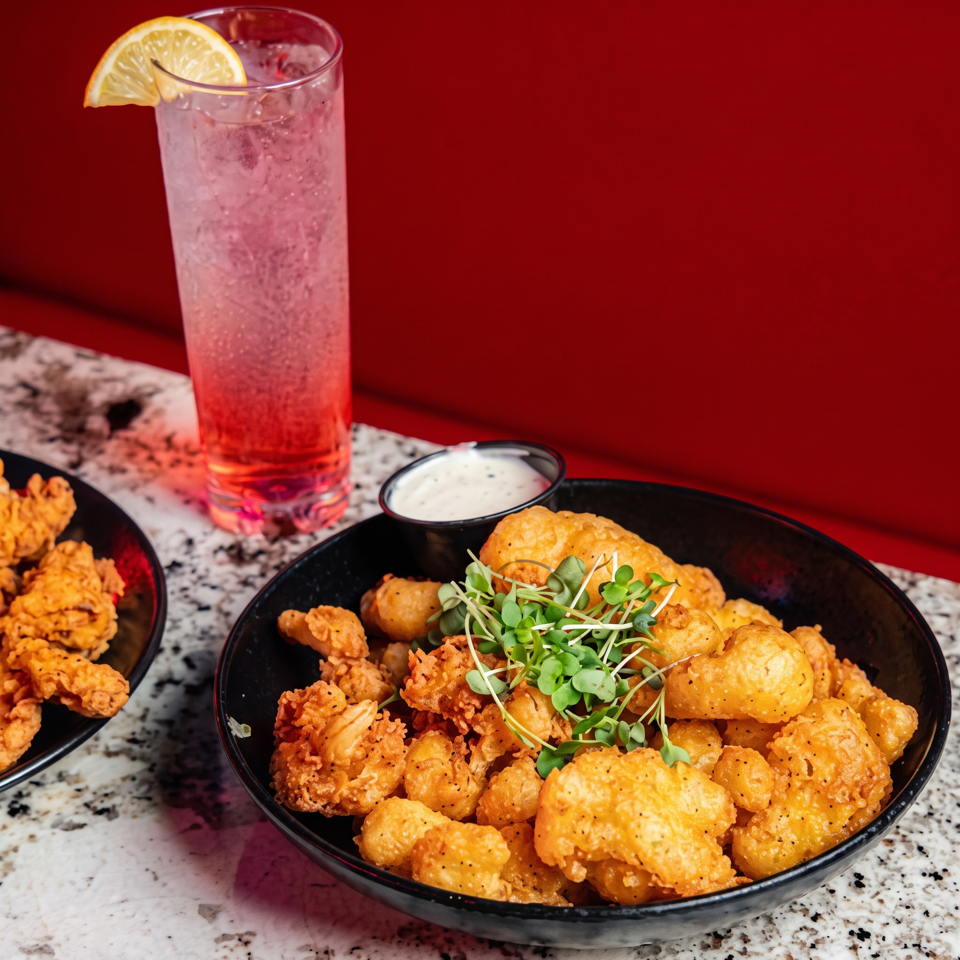 Black bowls of crispy fried bites with microgreens and dipping sauce beside a pink lemon-garnished drink on a speckled counter.
