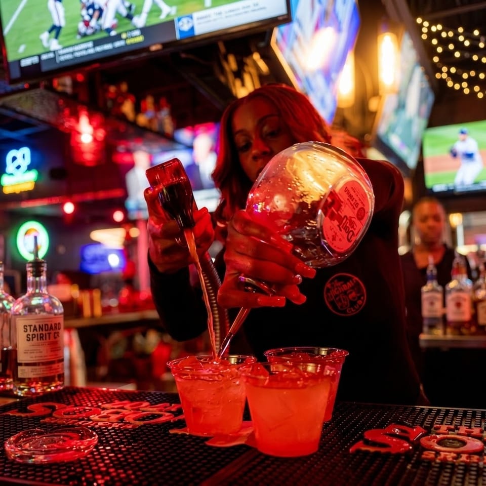 Bartender pours liquor into red cocktails at a lively sports bar surrounded by televisions and colorful lights.