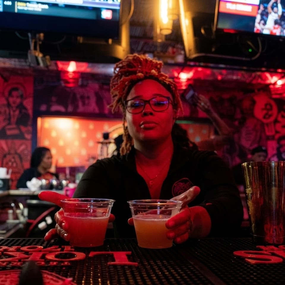 Bartender with dreadlocks presents two colorful cocktails at a red-lit bar surrounded by televisions and lively decor.